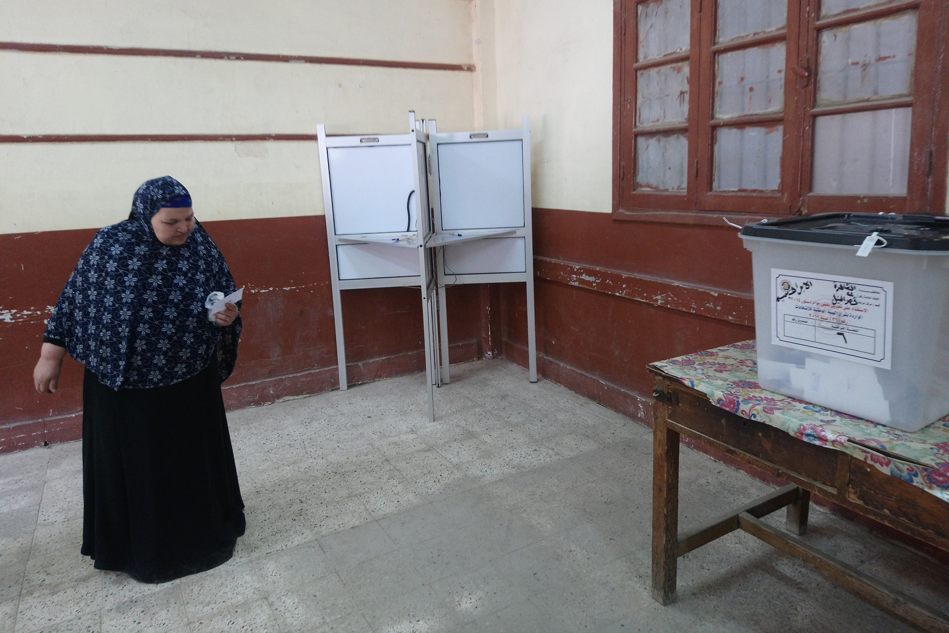 تُظهر الصورة امرأة داخل مركز اقتراع، وهي تحمل ورقة تصويت. وفي الخلفية، تظهر مقصورات التصويت وصندوق الاقتراع، مما يشير إلى أن الانتخابات جارية. ويحمل صندوق الاقتراع نصًا مكتوبًا باللغة العربية.