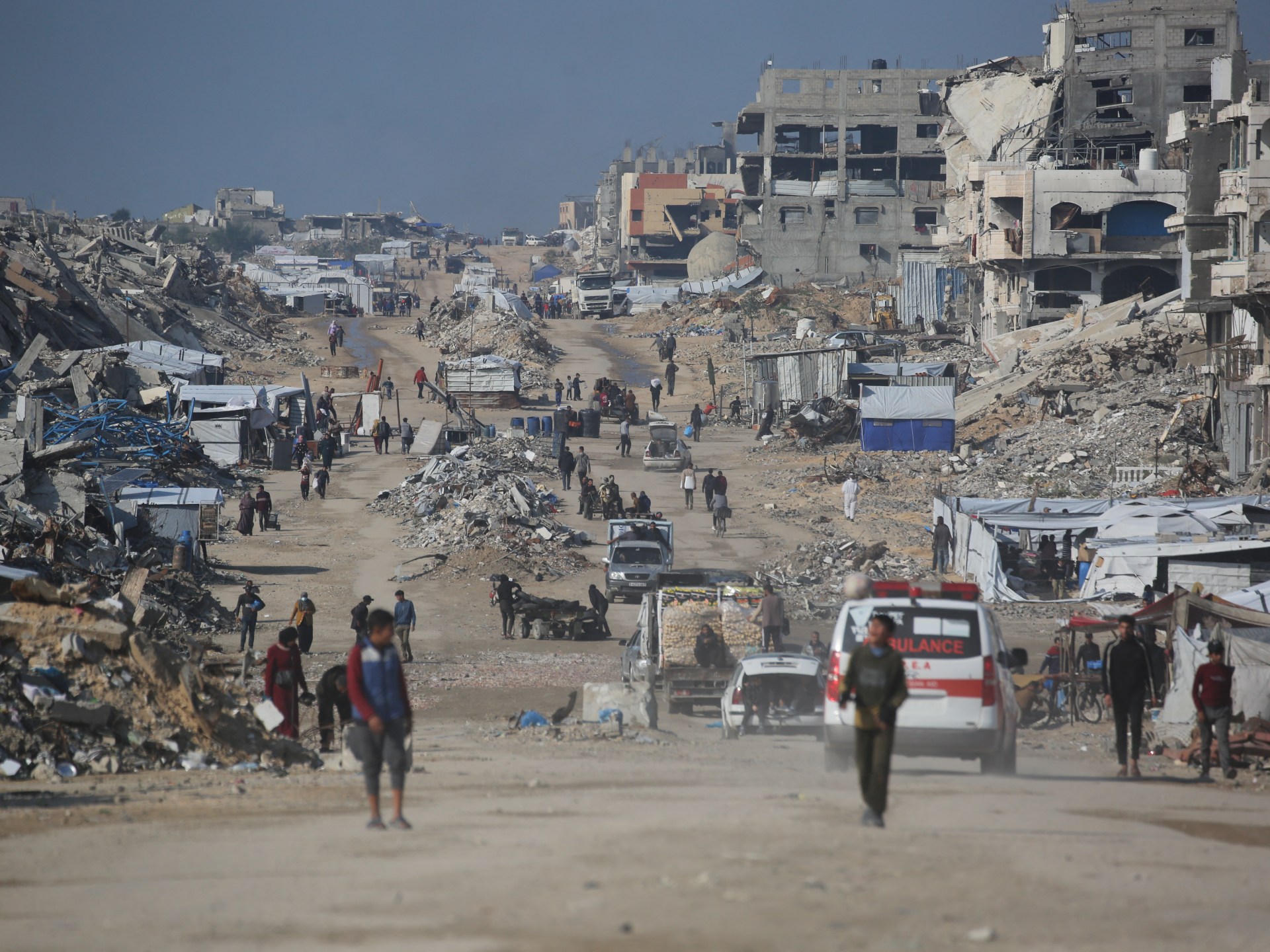 تصور الصورة مشهدًا لدمار هائل في غزة، حيث يسير الناس في شارع مليء بالأنقاض. تظهر سيارة إسعاف، مما يشير إلى استمرار الاستجابة الطارئة. تكشف الخلفية عن مبان متضررة بشدة. يوضح المشهد العام تداعيات الصراع، مما يعكس الأزمة الإنسانية الموصوفة في النص المقدم. يشير وجود المدنيين إلى النزوح والصمود وسط الخراب.